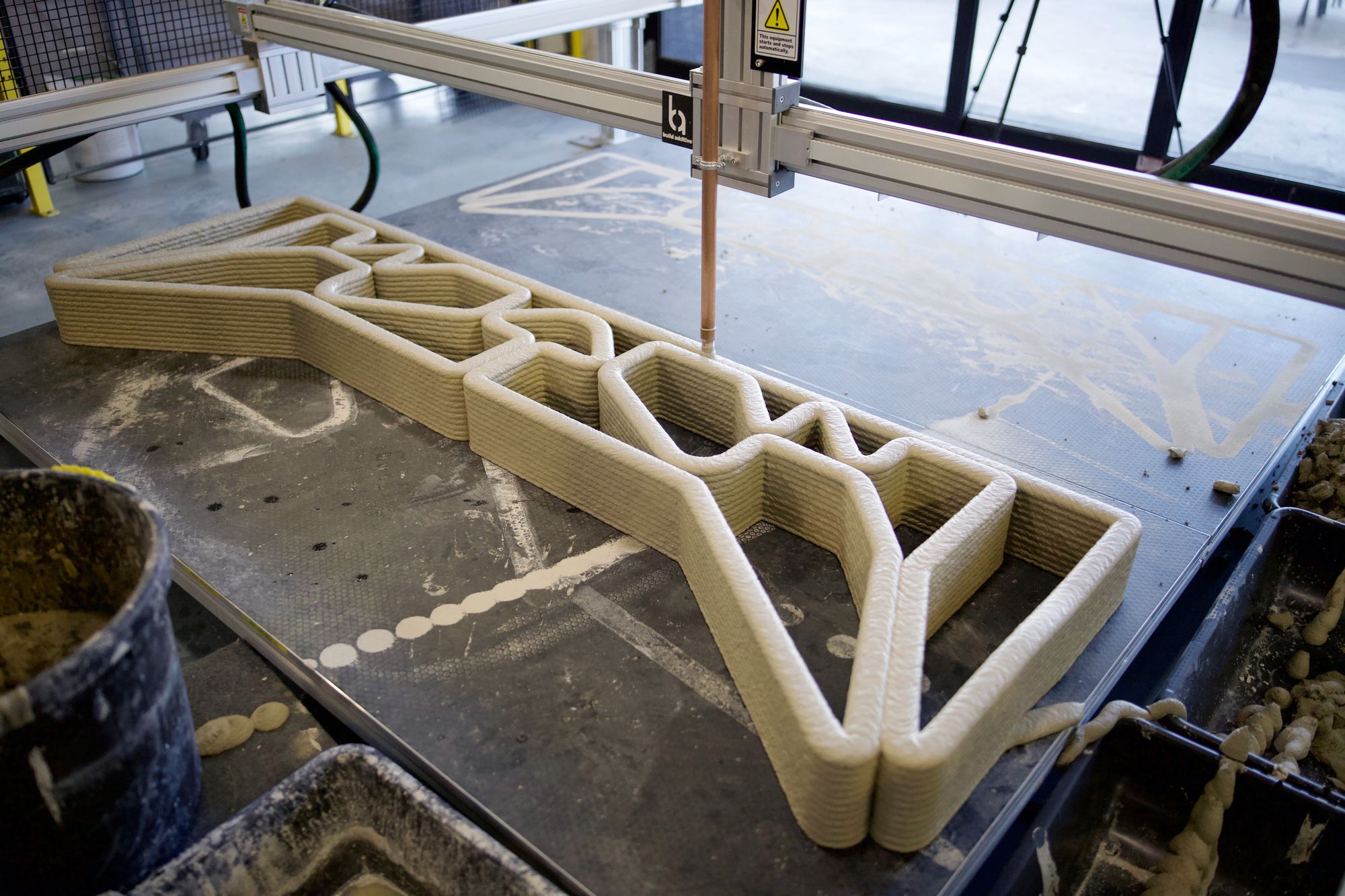 A large-scale 3D printer fabricating a light-colored concrete component with a geometric lattice structure on a flat build platform, surrounded by construction materials and equipment.